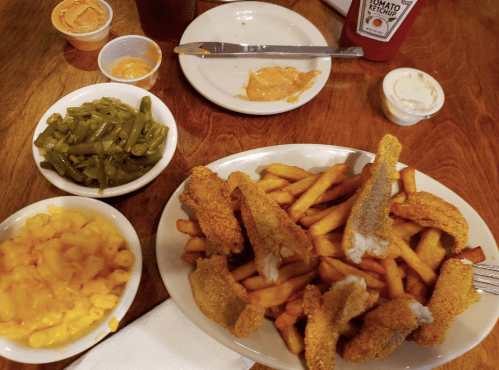 A plate of fried fish fillets on fries, with sides of macaroni and cheese, green beans, and condiments on a wooden table.