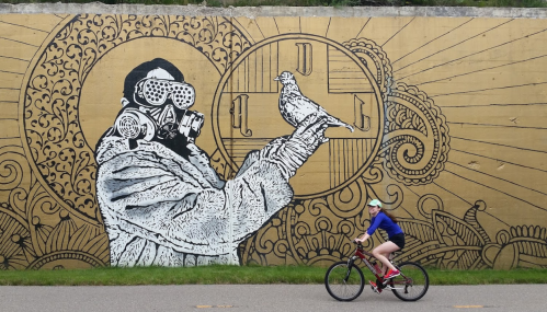 A cyclist rides past a large mural of a figure in a gas mask holding a bird, set against a golden background.