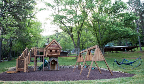 A wooden playground with a treehouse, swings, and a hammock, surrounded by lush green trees.