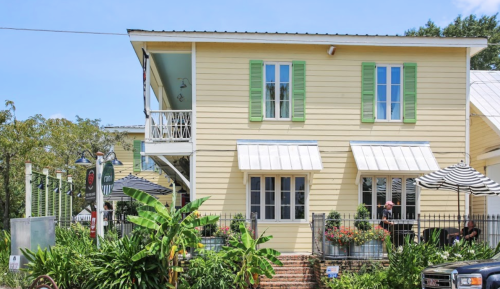 A charming yellow house with green shutters, featuring a balcony and surrounded by lush greenery and outdoor seating.
