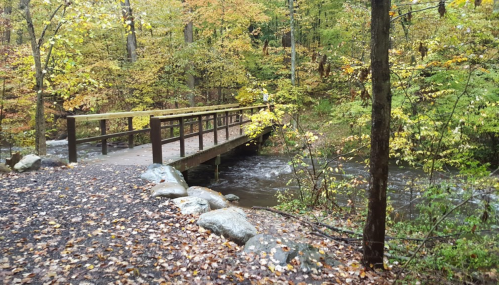 A wooden bridge over a flowing stream, surrounded by colorful autumn foliage and rocky terrain.