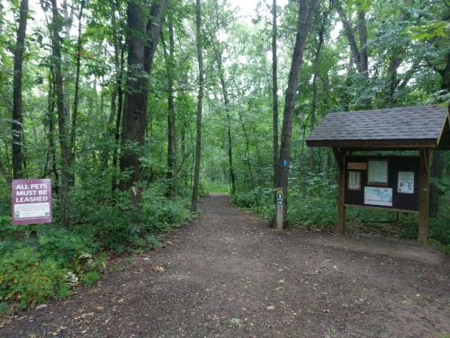 A wooded trail entrance with a sign about leashing pets and an information board under a shelter.