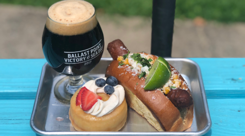 A glass of dark beer next to a hot dog topped with lime and a dessert donut with berries on a metal tray.