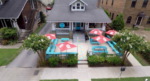 Aerial view of a café with colorful outdoor seating and red umbrellas, surrounded by greenery and nearby buildings.