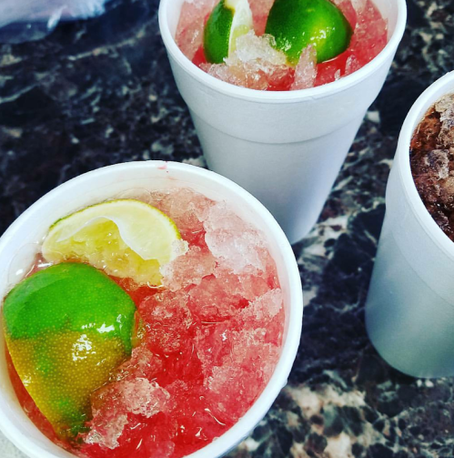 Two white cups filled with crushed ice, red drink, and lime and lemon slices on top, placed on a dark countertop.