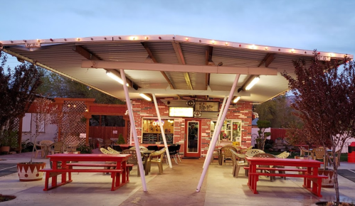 A colorful outdoor dining area with red picnic tables under a covered patio, surrounded by trees and lights.
