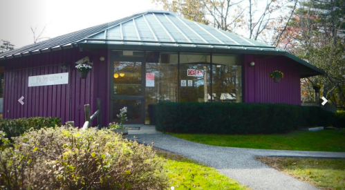 A purple building with a glass roof, featuring an "Open" sign, surrounded by greenery and a gravel path.