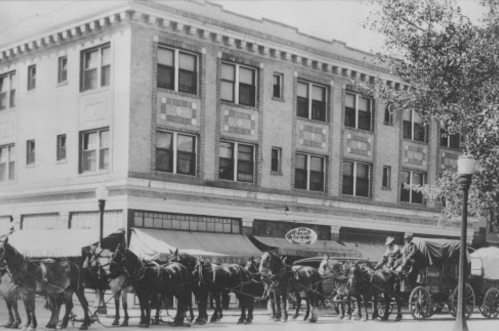 Historic black-and-white photo of horse-drawn carriages lined up in front of a brick building.
