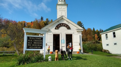 A charming chapel with a sign welcoming all breeds, featuring three dog statues in front against a colorful autumn backdrop.