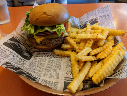 A cheeseburger with lettuce, tomato, and onion, served with crinkle-cut fries on a newspaper-lined plate.