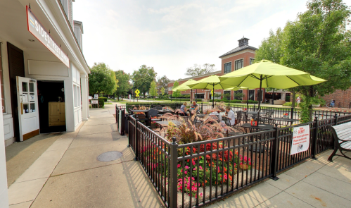 Outdoor dining area with green umbrellas, flowers, and patrons enjoying their meals near a building on a sunny day.