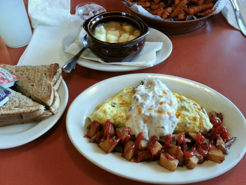 A plate with an omelet topped with gravy, diced potatoes with ketchup, toast, and a bowl of fruit on a red table.