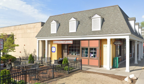 A quaint diner with a sloped roof, outdoor seating, and a sign reading "Sidestreet Diner" in a sunny setting.