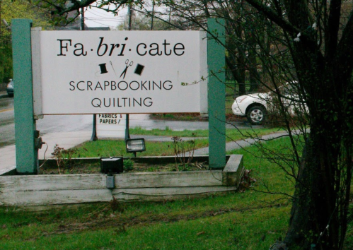 Sign for "Fa.bri.cate" featuring scrapbooking and quilting services, surrounded by greenery and a parked car.