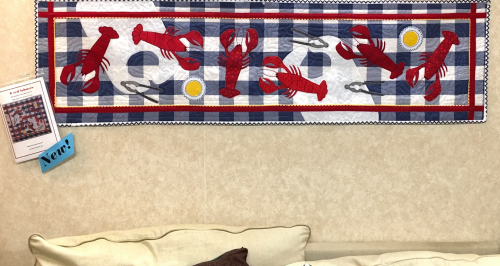 A quilt featuring red lobsters on a blue and white checkered background, displayed on a wall above a couch.