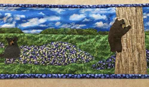 A colorful quilt featuring two bears, one climbing a tree and the other resting on a bed of purple flowers under a blue sky.