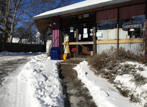 A cozy storefront with a "Welcome Open" sign, surrounded by snow and a clear path leading to the entrance.