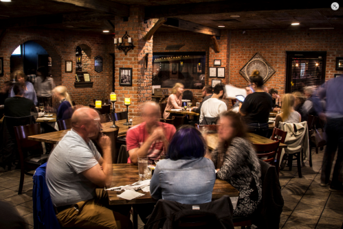 A bustling restaurant scene with blurred patrons at wooden tables, warm lighting, and brick walls creating a cozy atmosphere.