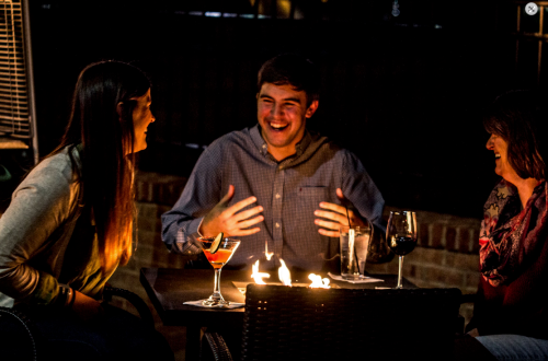 A group of three friends laughing and enjoying drinks around a table with a small fire at night.