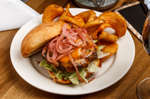 A juicy burger with melted cheese, lettuce, and pickled onions, served with crispy sweet potato fries on a plate.