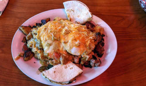 A plate of breakfast featuring two eggs, cheese, green sauce, potatoes, and tortillas on a wooden table.