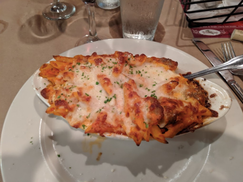 Baked pasta dish with melted cheese and herbs, served in a white dish on a plate, with utensils and drinks in the background.