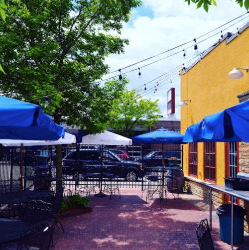 Outdoor dining area with blue umbrellas, string lights, and a vibrant yellow wall under a clear blue sky.