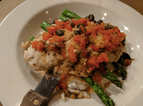 A plate of fish topped with diced tomatoes, olives, and capers, served on a bed of asparagus.
