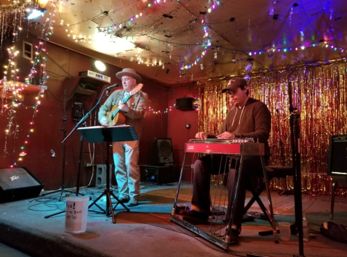 Two musicians perform on stage, one with a guitar and the other on a steel guitar, surrounded by colorful lights.