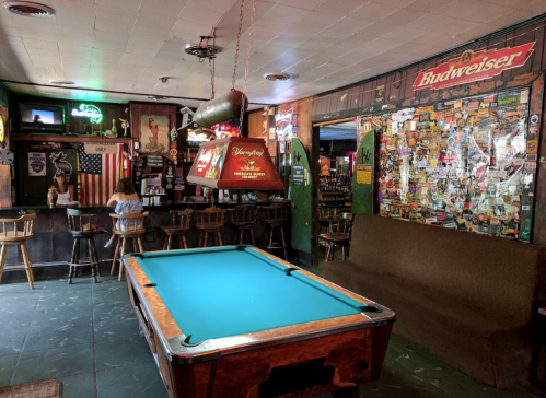 A cozy bar interior featuring a pool table, wooden stools, and a wall covered in stickers and memorabilia.
