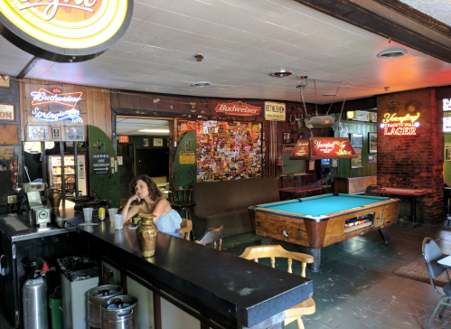 A cozy bar interior with a pool table, neon signs, and a woman sitting at the counter.