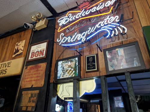 Neon Budweiser sign on a wooden wall, surrounded by framed photos and signs in a cozy bar setting.