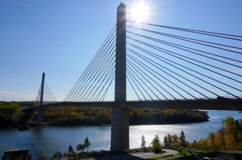 A modern cable-stayed bridge spans a river, with sunlight shining brightly above and autumn foliage in the background.