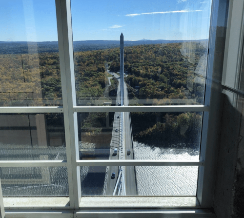 View of a bridge spanning a river, framed by large glass windows, with colorful autumn foliage in the background.