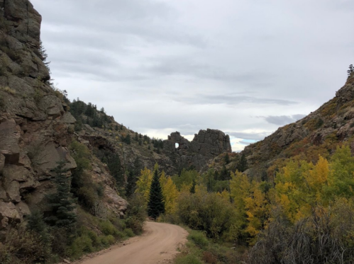 A dirt road winds through rocky terrain with trees and a distinctive rock formation in the distance under a cloudy sky.