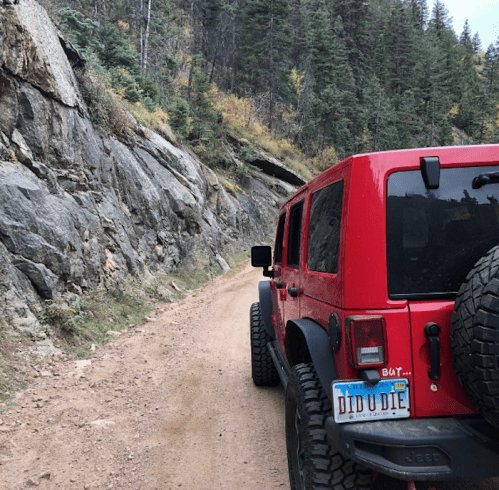 A red Jeep parked on a dirt road surrounded by rocky terrain and trees in a mountainous area.