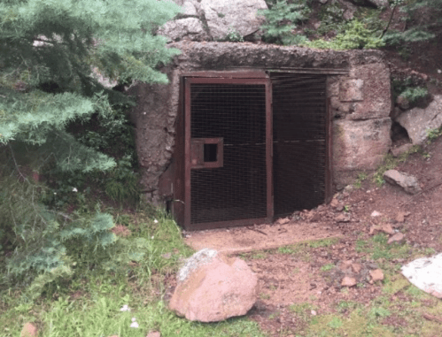 A weathered metal gate set into a rocky hillside, surrounded by greenery and scattered stones.