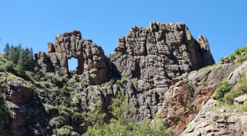 Rocky cliff formation with a natural arch, surrounded by greenery and a clear blue sky.