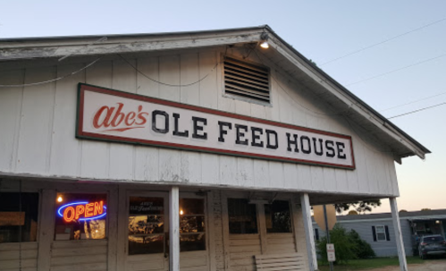 Sign for Abe's Ole Feed House, a rustic building with an "Open" neon sign, during twilight.