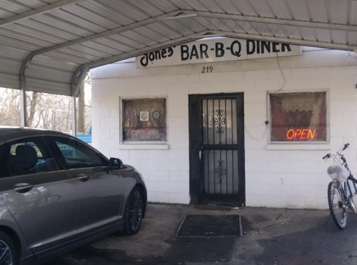 A small diner with a sign reading "Jones' Bar-B-Q Diner," featuring an open sign, a parked car, and a bicycle outside.