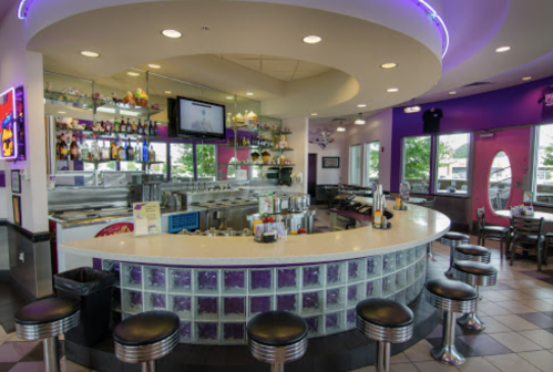 A modern diner interior featuring a curved bar, stools, and colorful lighting, with large windows and a vibrant atmosphere.