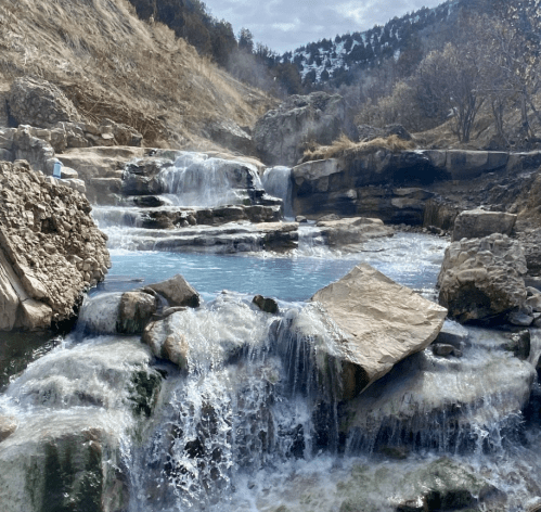 A serene waterfall cascades over rocks into a clear blue pool, surrounded by a rocky landscape and distant trees.