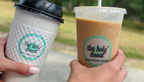 Two hands hold a coffee cup and an iced drink, both featuring "The Holy Donut" logo.