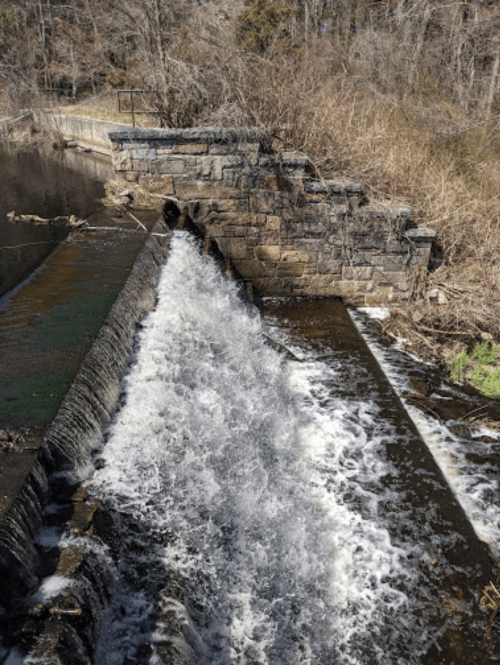 Water cascades over a stone dam, surrounded by bare trees and brush, under a clear blue sky.