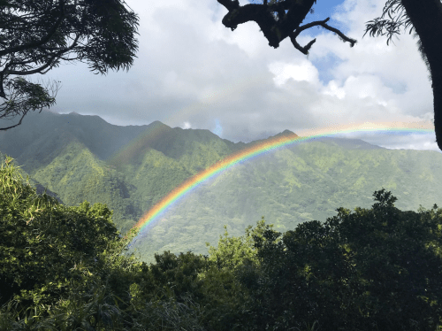 A vibrant rainbow arcs over lush green mountains under a partly cloudy sky.
