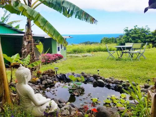 A serene garden with a Buddha statue, pond, and ocean view, surrounded by lush greenery and colorful flowers.