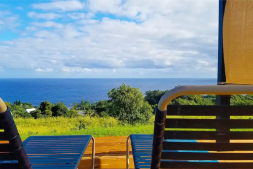 Two lounge chairs overlook a serene ocean view under a partly cloudy sky. Lush greenery surrounds the area.