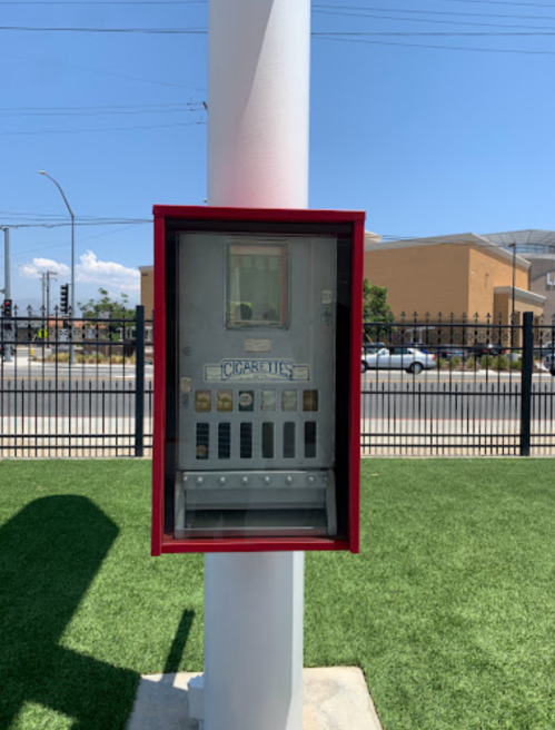A red cigarette vending machine mounted on a white pole, set against a grassy area and blue sky.