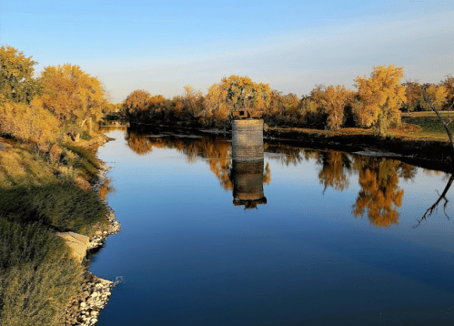 A calm river reflects autumn trees and a stone pillar, with clear blue skies above.