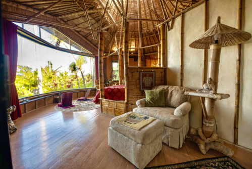 Cozy interior of a bamboo-style room with a bed, armchair, and large windows showcasing lush greenery outside.